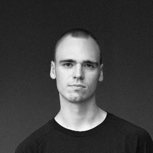 Black and white portrait of a young man with short hair wearing a dark shirt against a plain dark background.