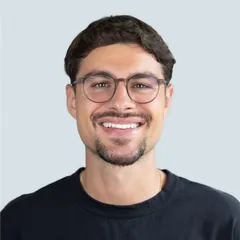 Smiling young man with glasses, short dark hair, and a goatee wearing a black shirt against a light background.