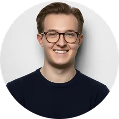 Smiling young man wearing glasses and a dark sweater against a light gray background.